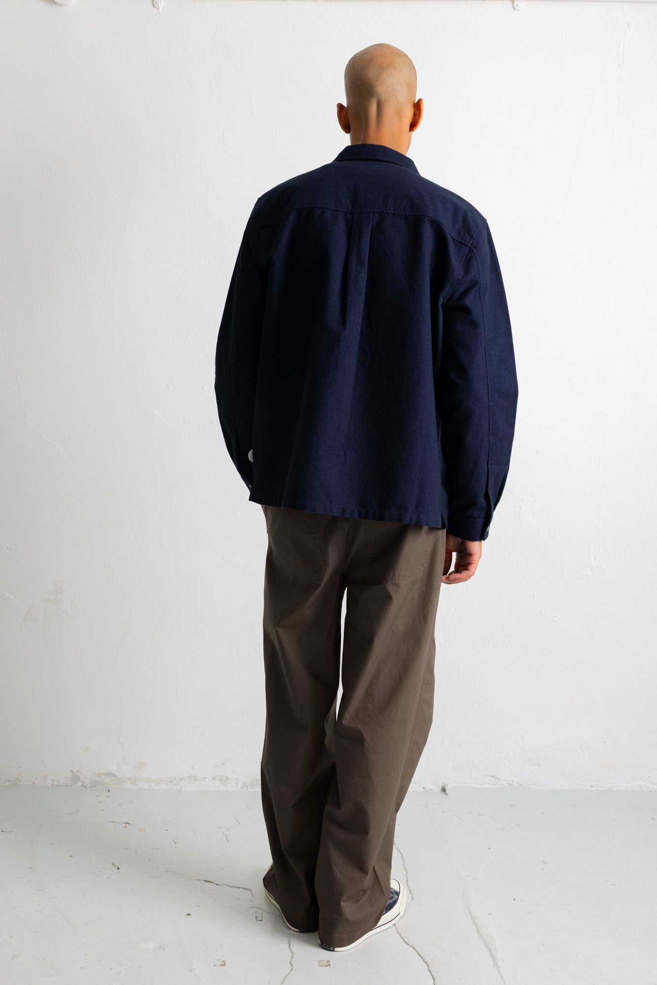 A man viewed from behind wearing a dark navy blue long-sleeve button-up shirt and brown trousers, standing against a plain white wall.