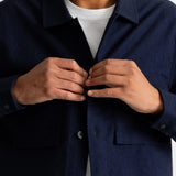 A close-up of a man wearing a dark navy blue overshirt over a white t-shirt, with his hands buttoning the collar of the overshirt.