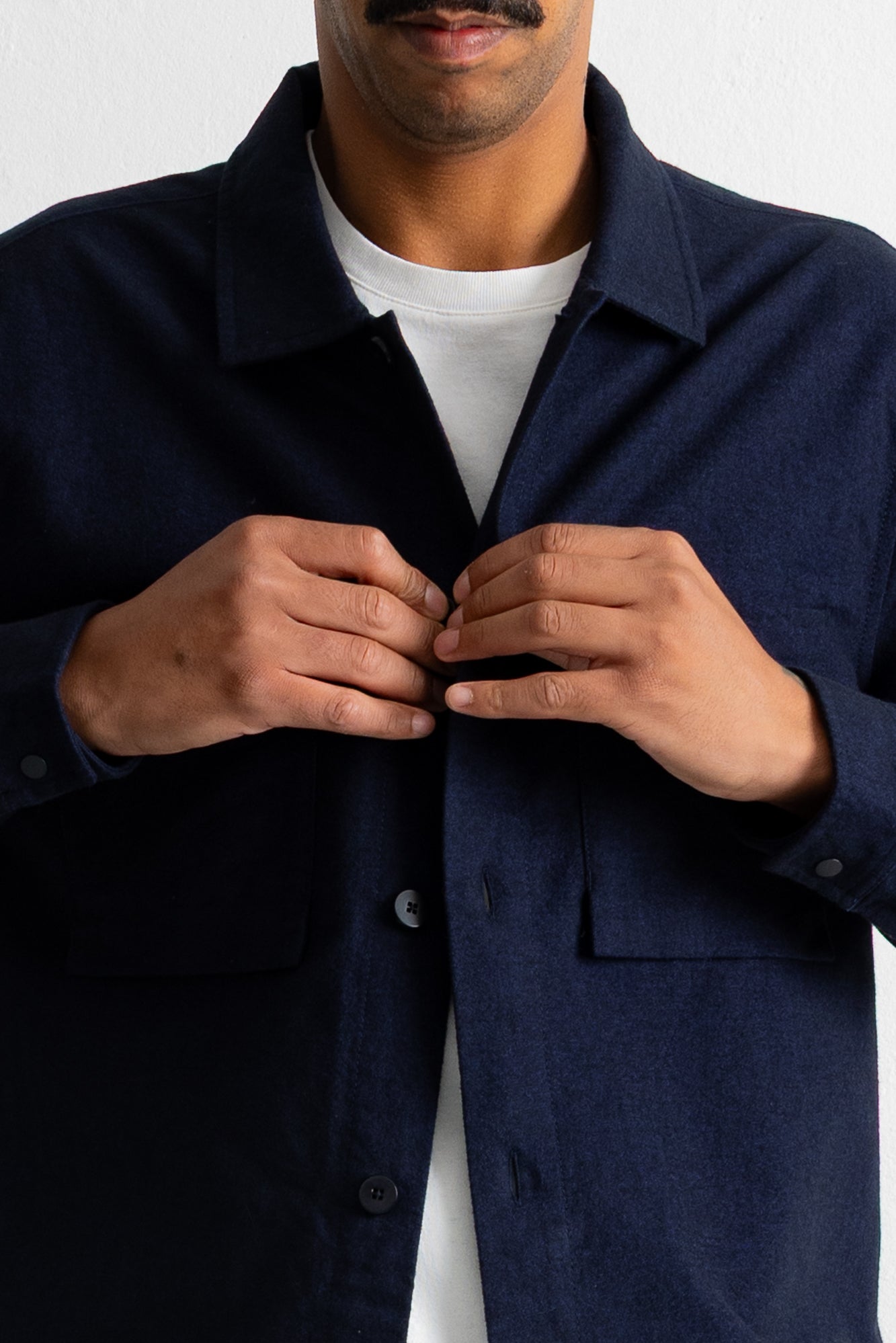 A close-up of a man wearing a dark navy blue overshirt over a white t-shirt, with his hands buttoning the collar of the overshirt.
