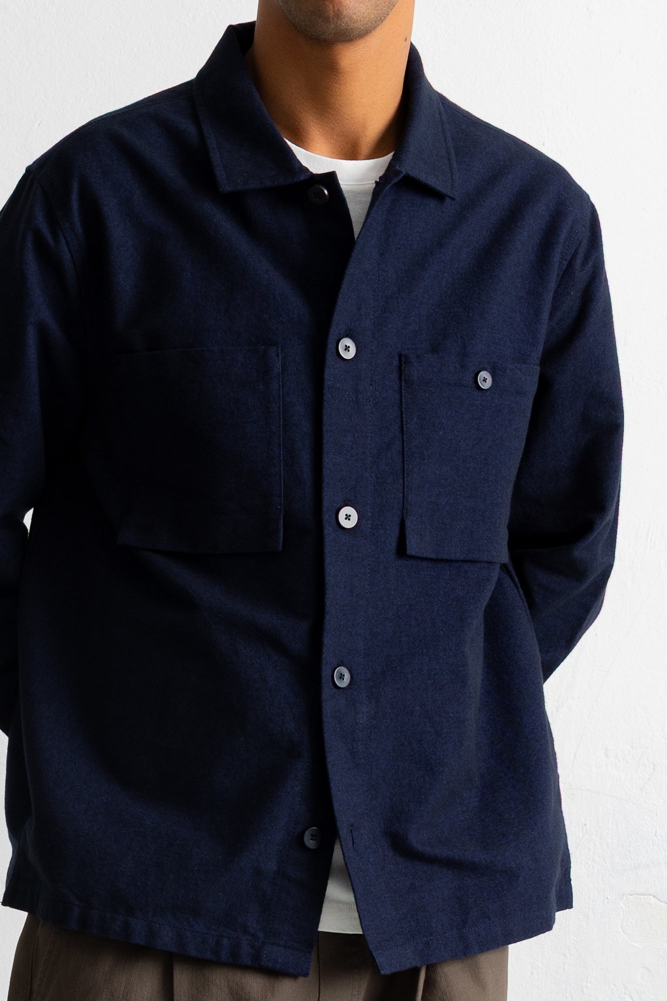 A man wearing a dark navy blue long-sleeve button-up overshirt over a white t-shirt, shown from the neck down against a white wall.