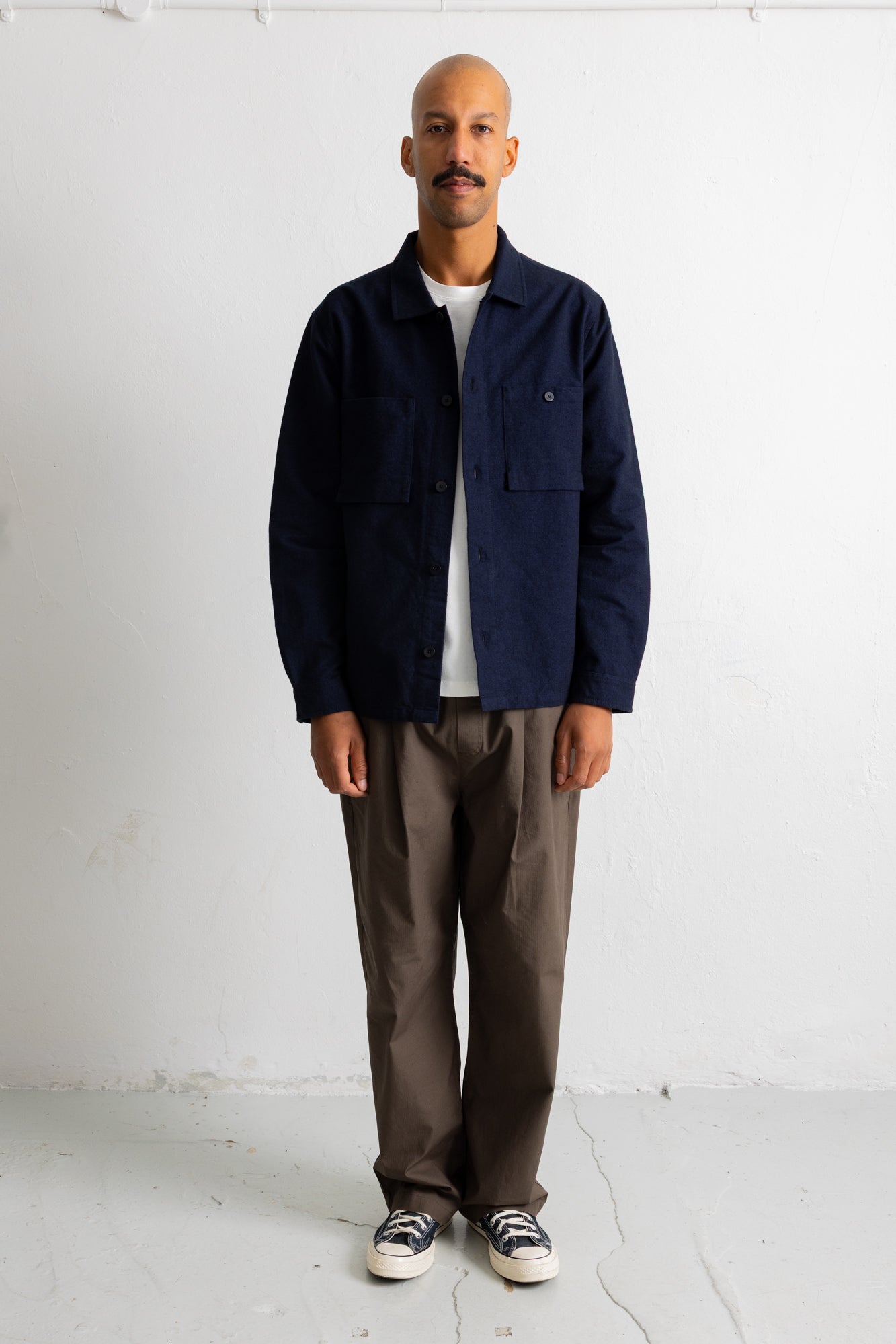 A man stands against a white wall wearing a dark navy blue utility overshirt over a white t-shirt, paired with brown pants and black-and-white sneakers.
