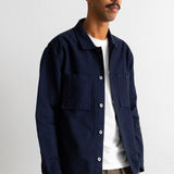 A man wearing a dark navy blue button-up overshirt over a white t-shirt and brown pants, standing against a plain off-white wall.