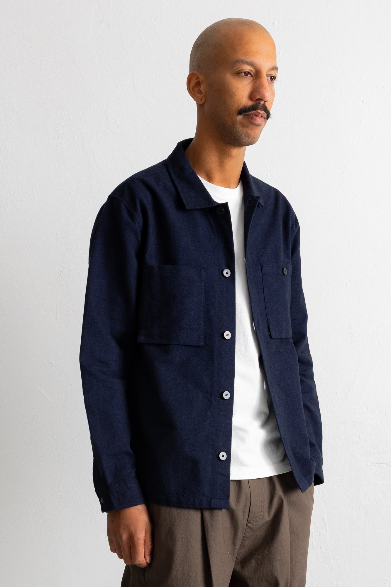 A man wearing a dark navy blue button-up overshirt over a white t-shirt and brown pants, standing against a plain off-white wall.