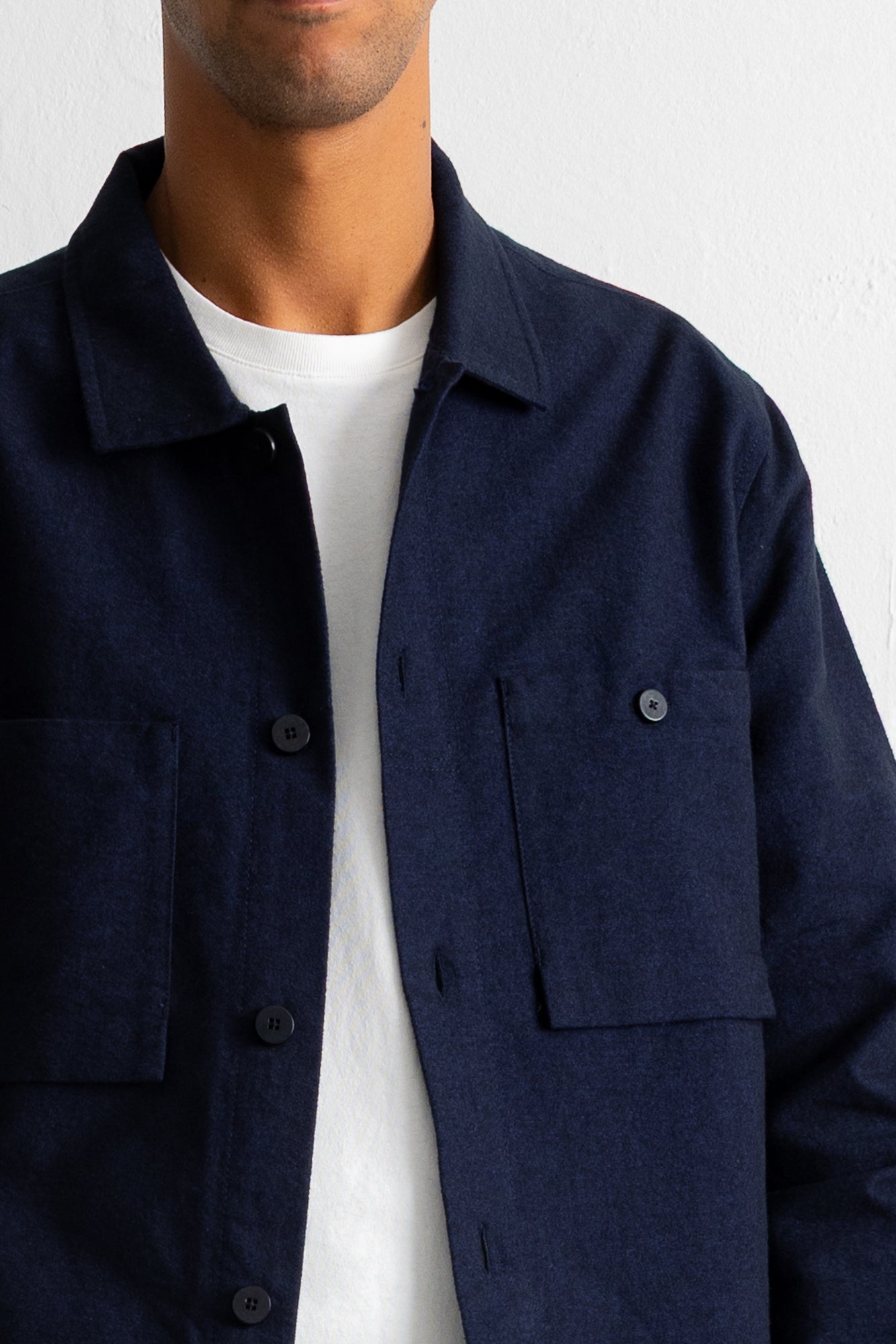 A close-up of a person wearing a dark navy blue button-up overshirt over a white t-shirt, against a plain white wall.