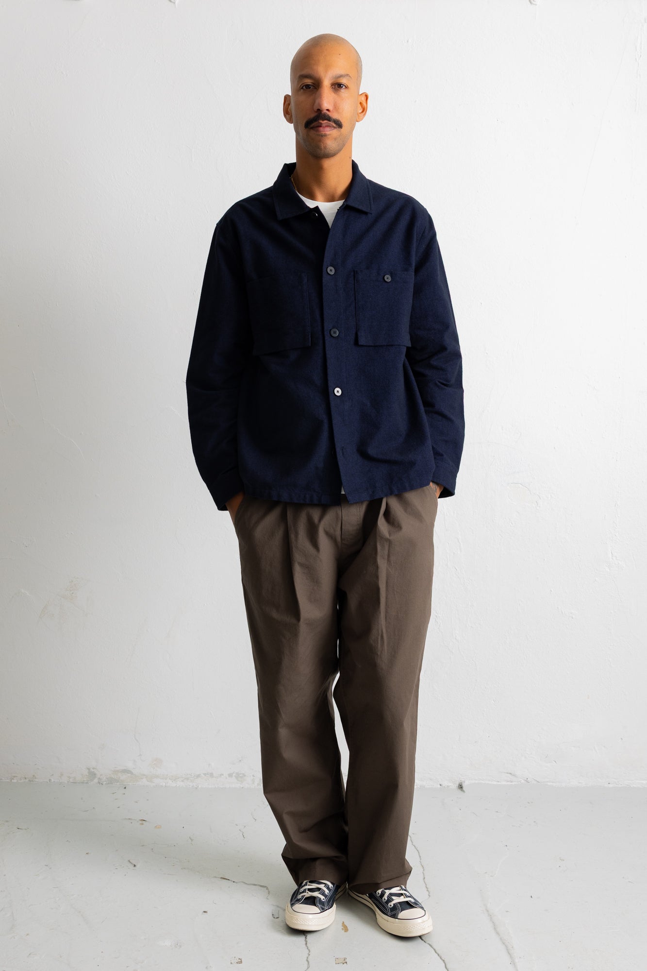 A man wearing a dark navy blue button-up overshirt, brown wide-leg trousers, and black-and-white sneakers, standing against a plain white studio wall.