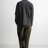 A man viewed from behind wearing a dark gray and black plaid button-down shirt, olive green corduroy pants, and black canvas sneakers, standing against a white wall.
