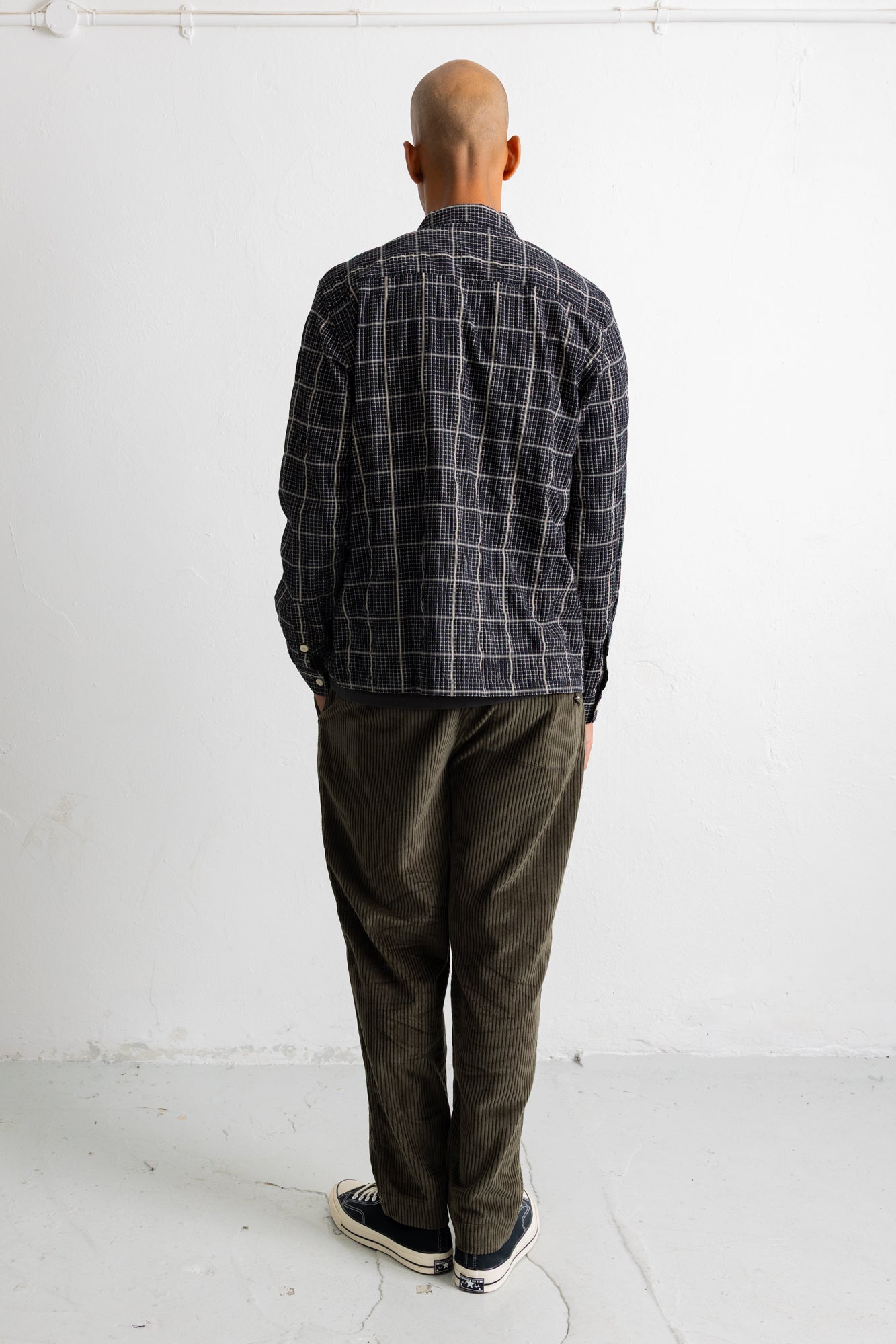 A man viewed from behind wearing a dark gray and black plaid button-down shirt, olive green corduroy pants, and black canvas sneakers, standing against a white wall.