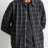 A man wearing a black and white plaid overshirt over a dark gray t-shirt and brown corduroy trousers, shown from the chin down against a plain light gray background.