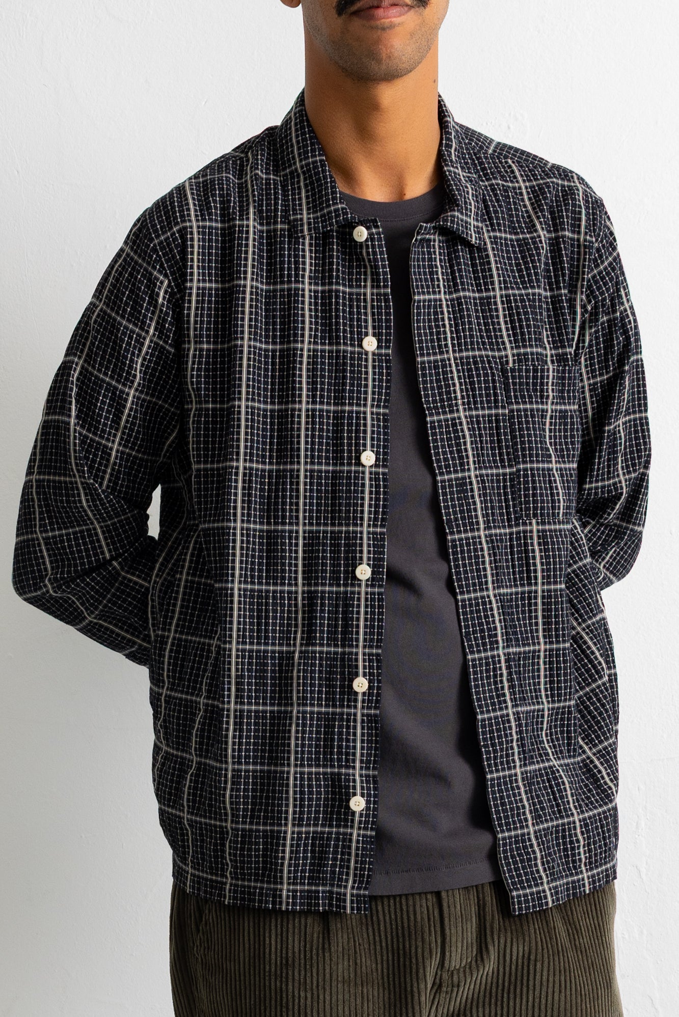A man wearing a black and white plaid overshirt over a dark gray t-shirt and brown corduroy trousers, shown from the chin down against a plain light gray background.