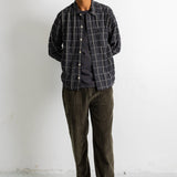 A man wearing a dark gray and black plaid overshirt over a dark t-shirt, dark olive corduroy pants, and black canvas sneakers, standing against a white wall.