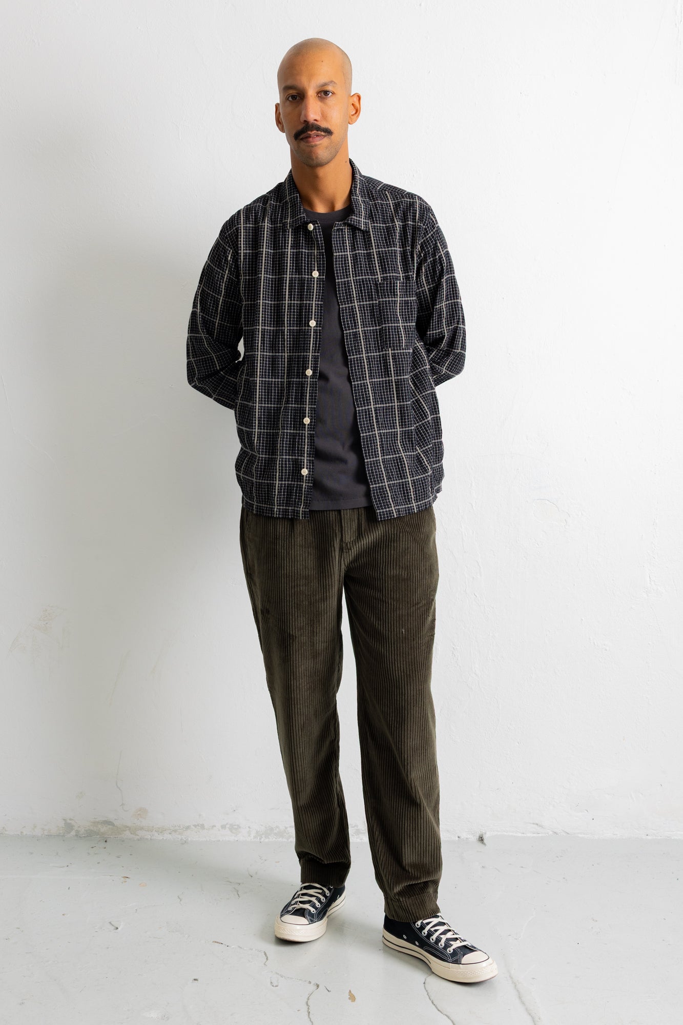 A man wearing a dark gray and black plaid overshirt over a dark t-shirt, dark olive corduroy pants, and black canvas sneakers, standing against a white wall.