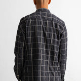 A man viewed from behind wearing a dark navy and white windowpane plaid button-down shirt and dark olive corduroy trousers, standing against a plain white wall.