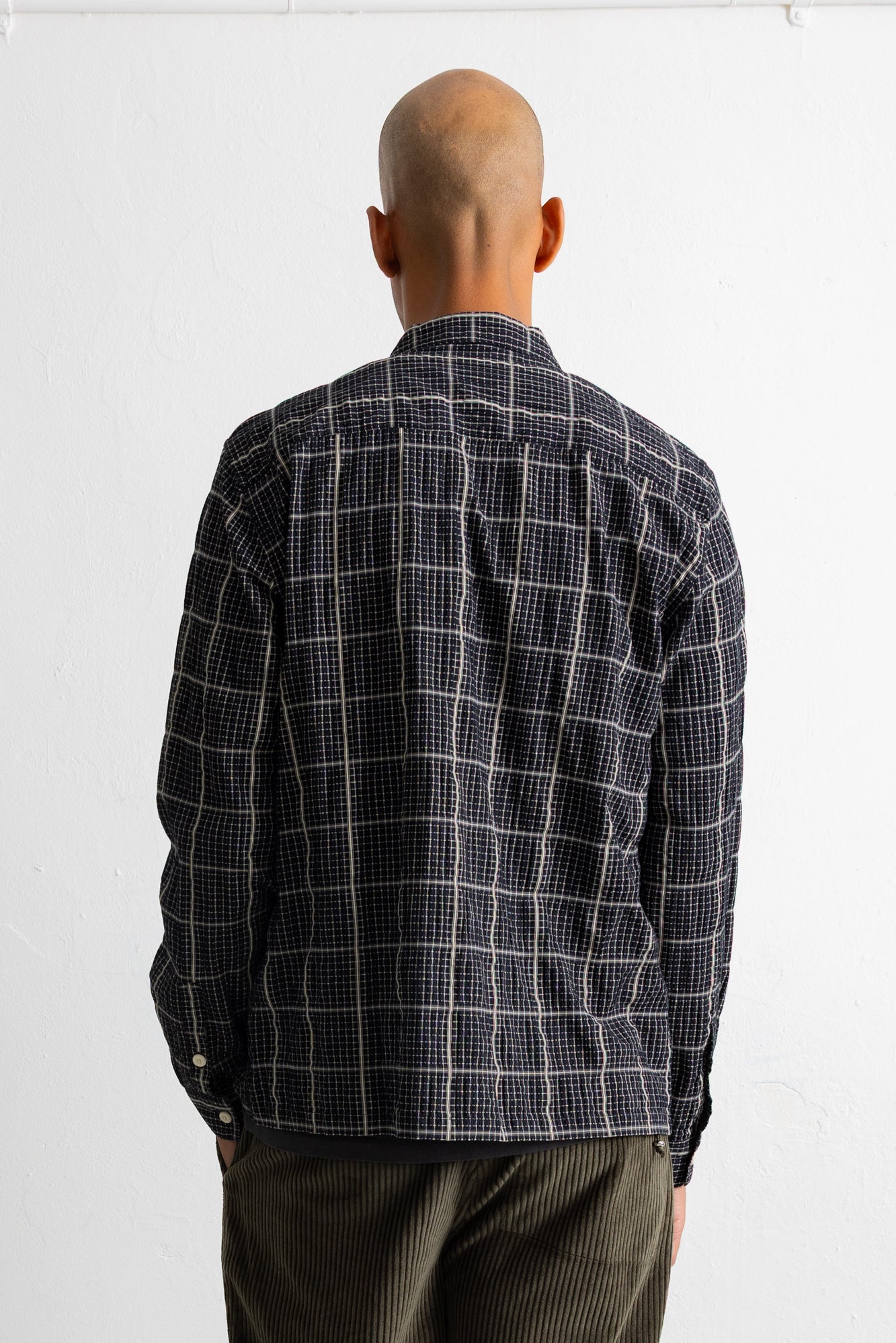 A man viewed from behind wearing a dark navy and white windowpane plaid button-down shirt and dark olive corduroy trousers, standing against a plain white wall.