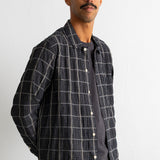 A man wearing a dark gray and black plaid overshirt over a dark gray t-shirt and brown corduroy pants, standing against a white wall.