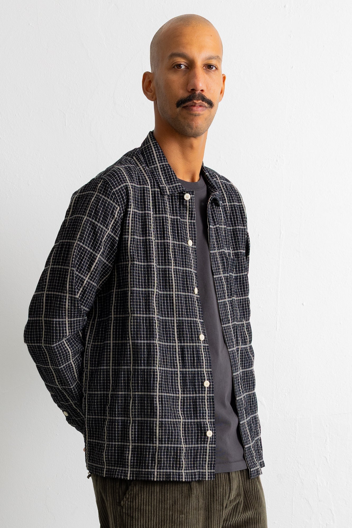 A man wearing a dark gray and black plaid overshirt over a dark gray t-shirt and brown corduroy pants, standing against a white wall.