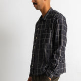A man wearing a dark navy and black plaid button-down shirt with white grid lines, paired with olive green corduroy trousers, standing against a plain white wall.