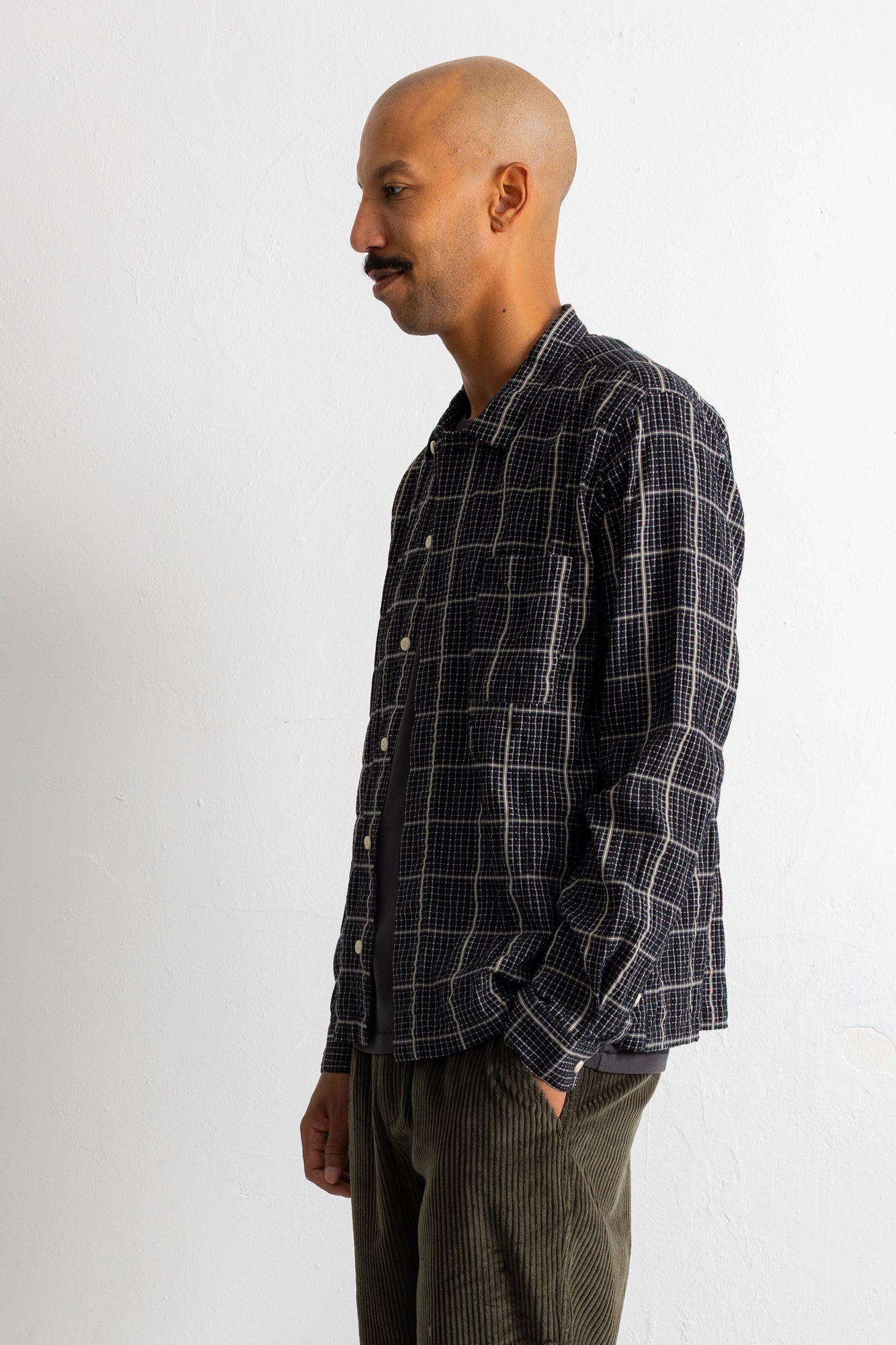 A man wearing a dark navy and black plaid button-down shirt with white grid lines, paired with olive green corduroy trousers, standing against a plain white wall.