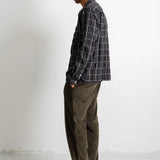 A man stands in profile wearing a dark gray and black plaid button-down shirt, olive green corduroy pants, and black canvas sneakers against a plain white wall.