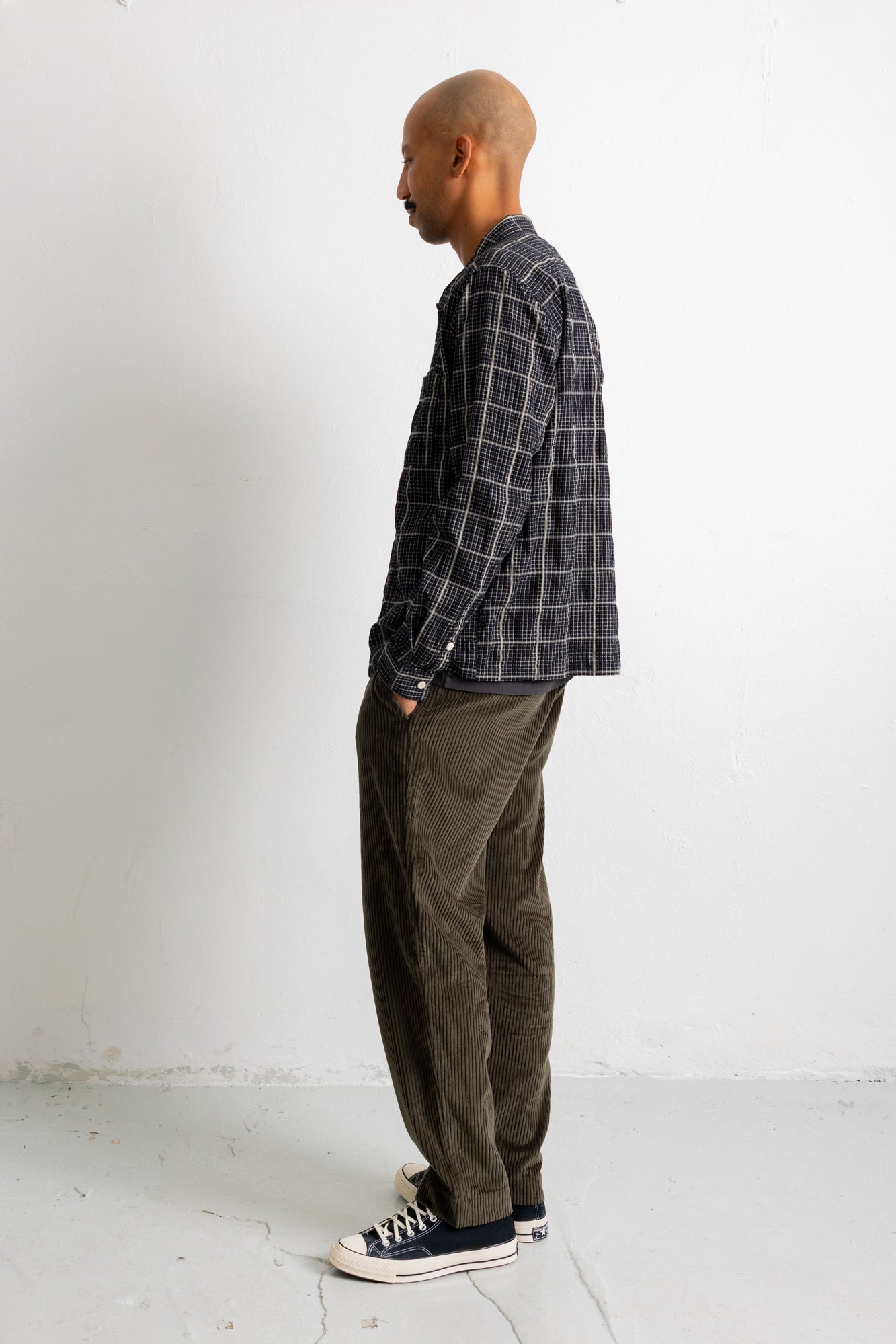 A man stands in profile wearing a dark gray and black plaid button-down shirt, olive green corduroy pants, and black canvas sneakers against a plain white wall.