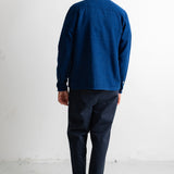 A man viewed from behind wearing a dark blue long-sleeve button-up shirt, dark navy pants, and black canvas sneakers, standing against a plain white wall.