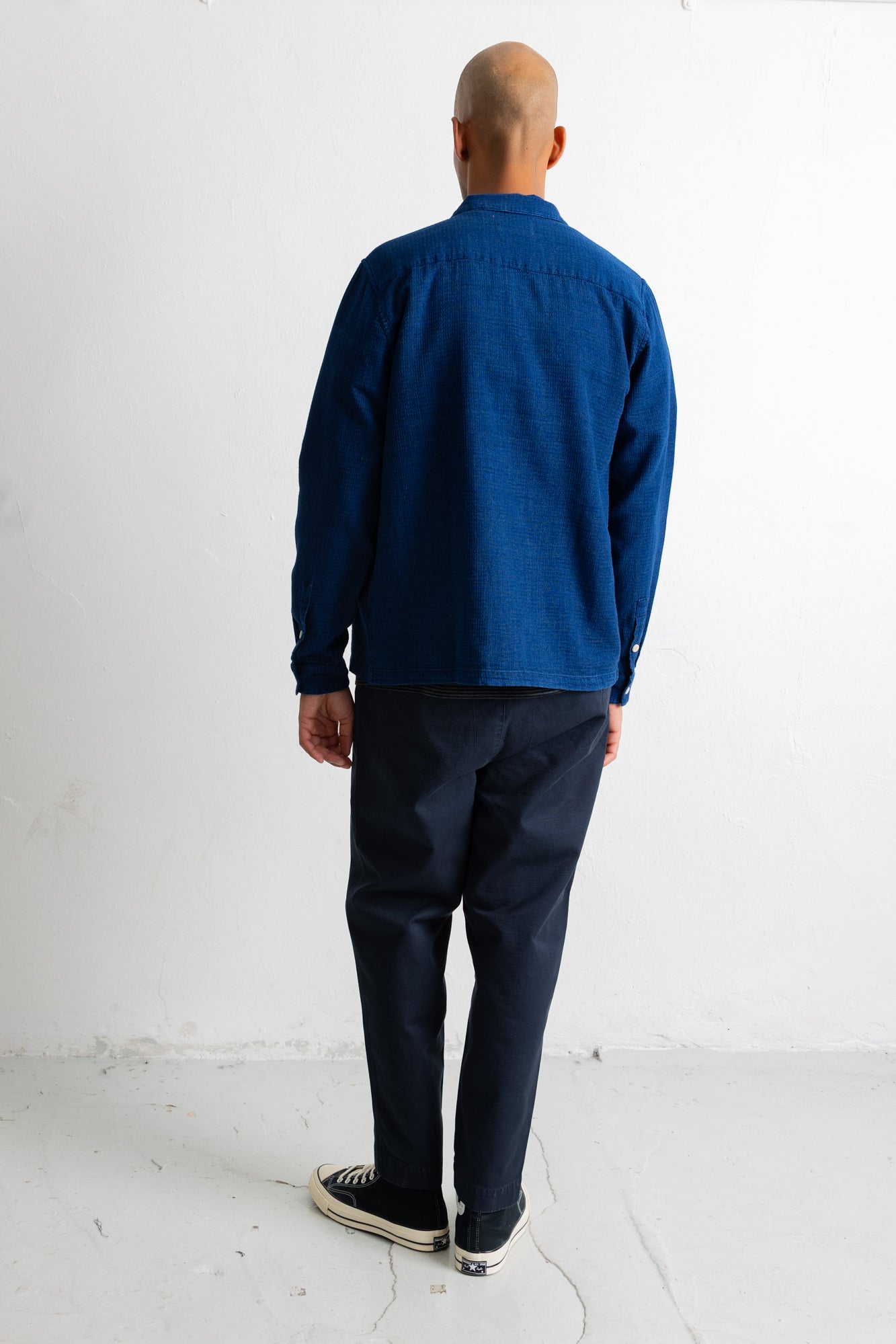 A man viewed from behind wearing a dark blue long-sleeve button-up shirt, dark navy pants, and black canvas sneakers, standing against a plain white wall.