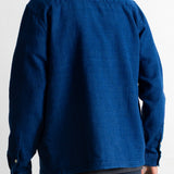 A man viewed from the back wearing a textured royal blue long-sleeve button-up shirt and dark trousers, set against a plain white background.