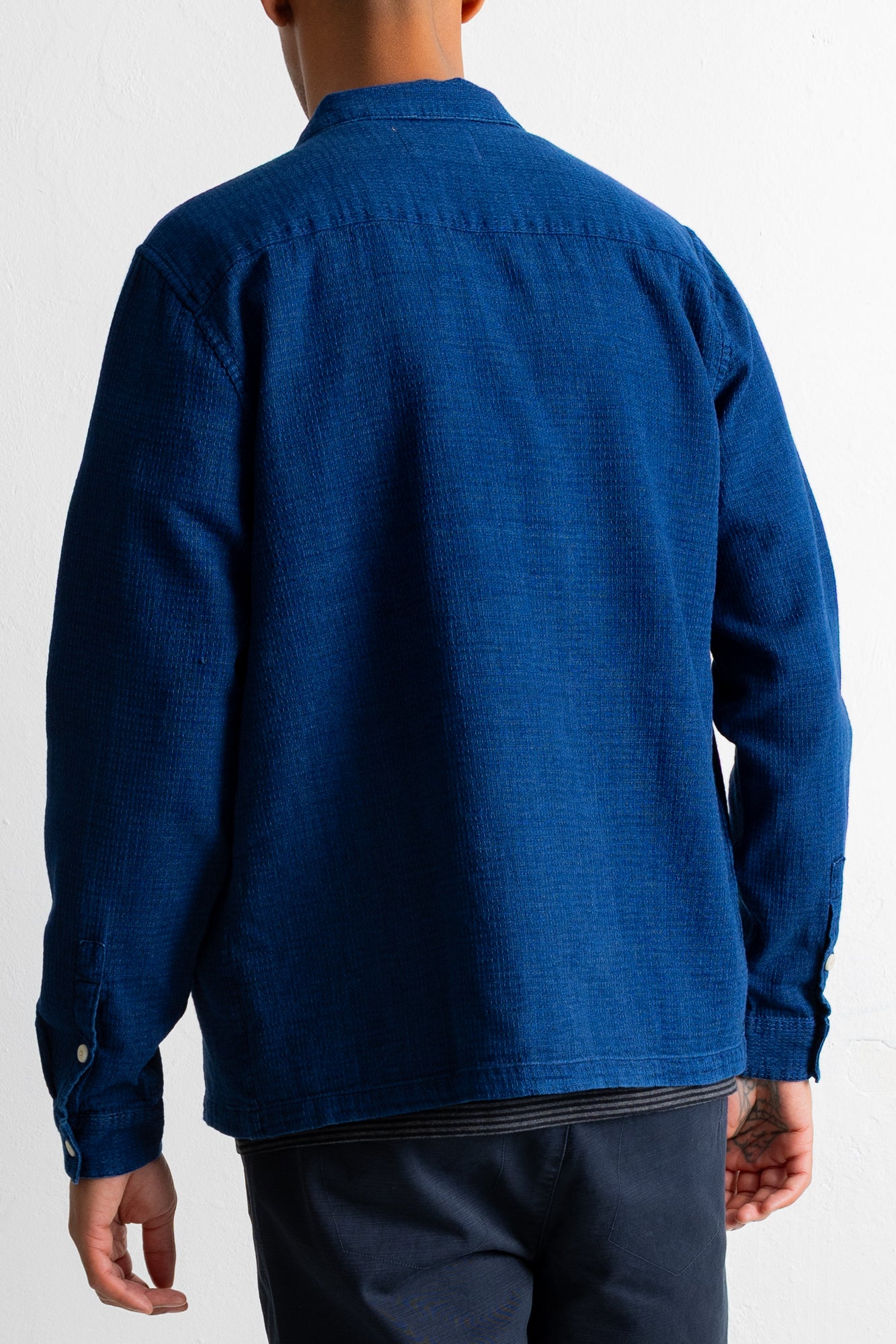 A man viewed from the back wearing a textured royal blue long-sleeve button-up shirt and dark trousers, set against a plain white background.