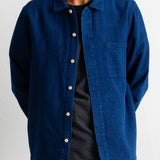 A man wearing a textured blue button-up overshirt over a black striped t-shirt and navy pants, shown from the chin down against a white wall.