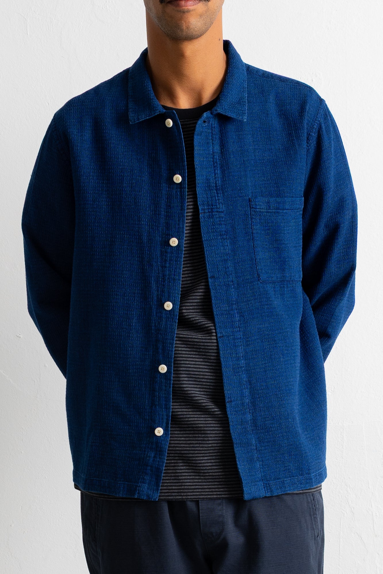 A man wearing a textured blue button-up overshirt over a black striped t-shirt and navy pants, shown from the chin down against a white wall.