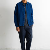 A man wearing a blue button-up overshirt over a dark gray t-shirt, dark navy pants, and black-and-white sneakers, standing against a white wall.