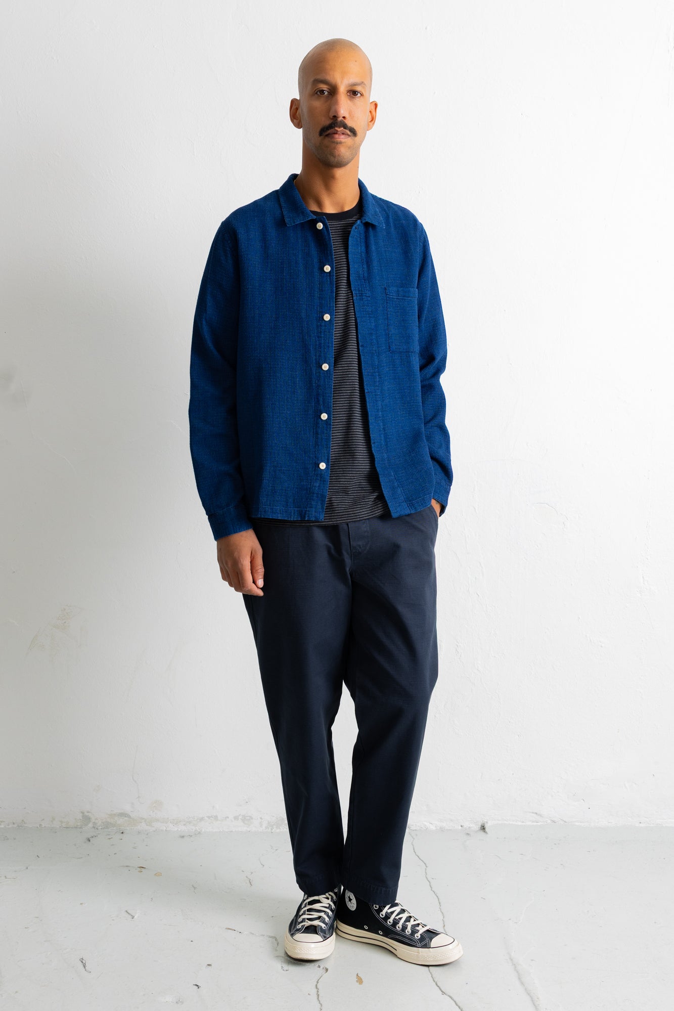 A man wearing a blue button-up overshirt over a dark gray t-shirt, dark navy pants, and black-and-white sneakers, standing against a white wall.