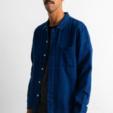 A man wearing a dark indigo blue long-sleeve button-up shirt over a black striped t-shirt and dark trousers, standing against a plain white wall.