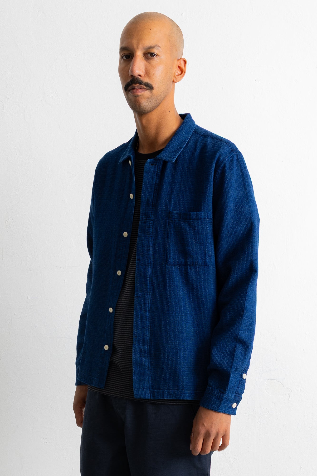 A man wearing a dark indigo blue long-sleeve button-up shirt over a black striped t-shirt and dark trousers, standing against a plain white wall.