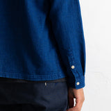 Close-up of a person wearing a textured indigo blue long-sleeve shirt with dark navy pants, showing the sleeve button detail against a white wall.