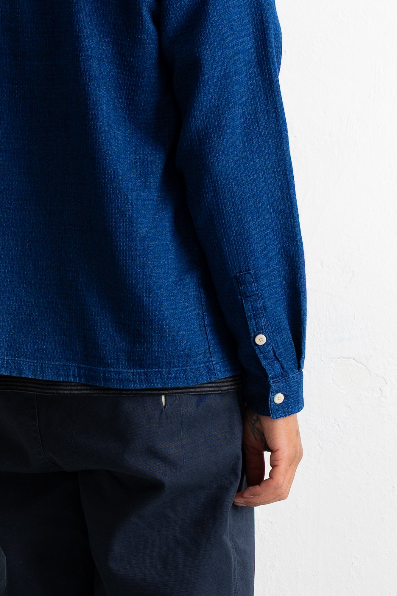 Close-up of a person wearing a textured indigo blue long-sleeve shirt with dark navy pants, showing the sleeve button detail against a white wall.