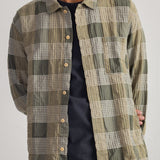 A man wearing a long-sleeve button-up plaid overshirt in olive green, beige, and white, layered over a black t-shirt and blue jeans.