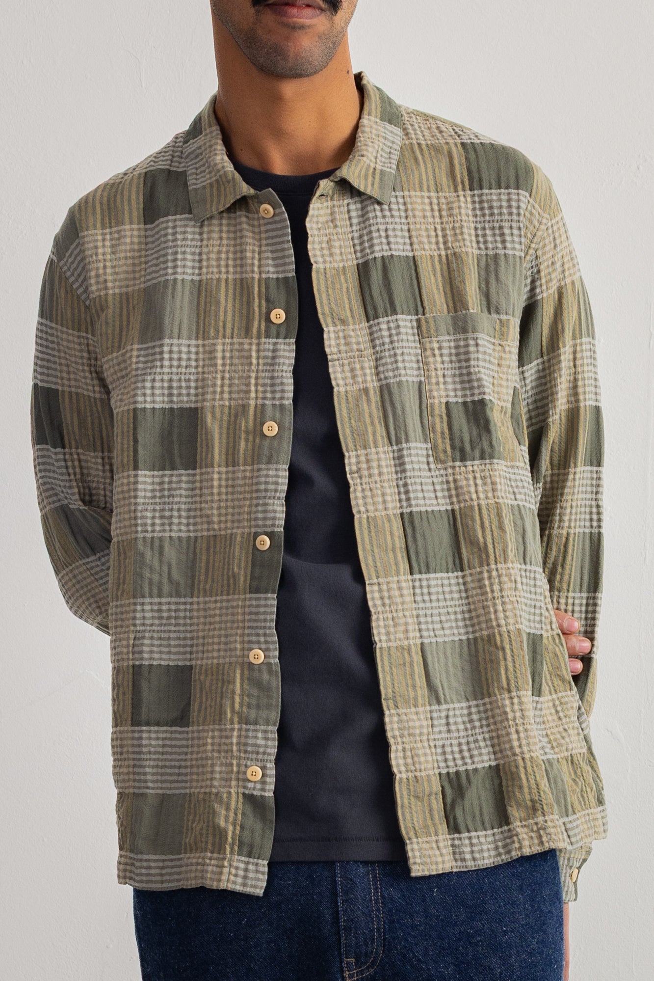 A man wearing a long-sleeve button-up plaid overshirt in olive green, beige, and white, layered over a black t-shirt and blue jeans.