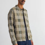 A man wearing a long-sleeve button-up shirt with a muted green and beige plaid pattern, layered over a dark gray t-shirt and blue jeans, standing against a white wall.