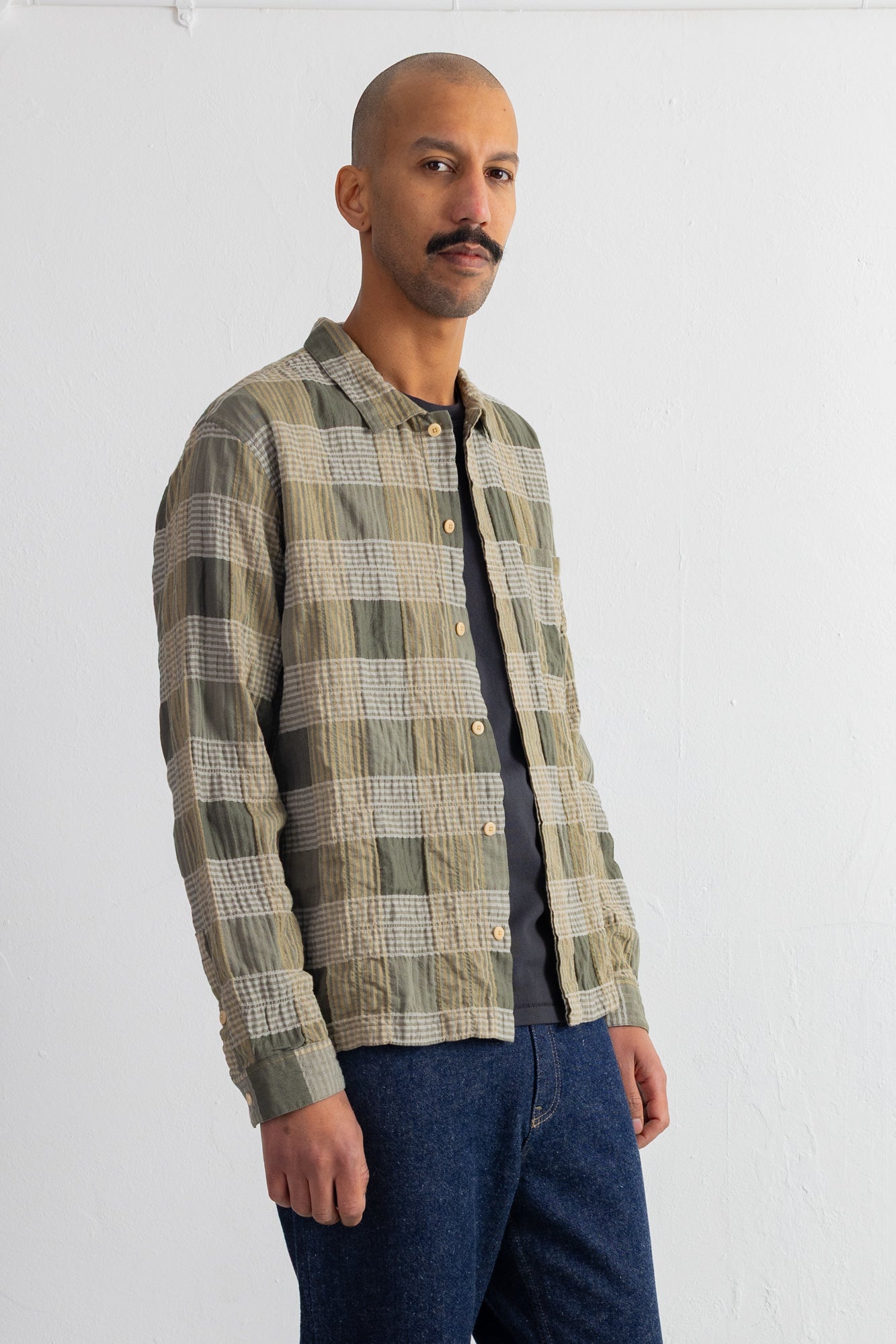 A man wearing a long-sleeve button-up shirt with a muted green and beige plaid pattern, layered over a dark gray t-shirt and blue jeans, standing against a white wall.