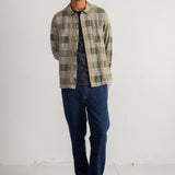 A man wearing a green and beige plaid button-up shirt over a dark t-shirt, paired with dark blue jeans and black shoes, standing against a white studio wall.