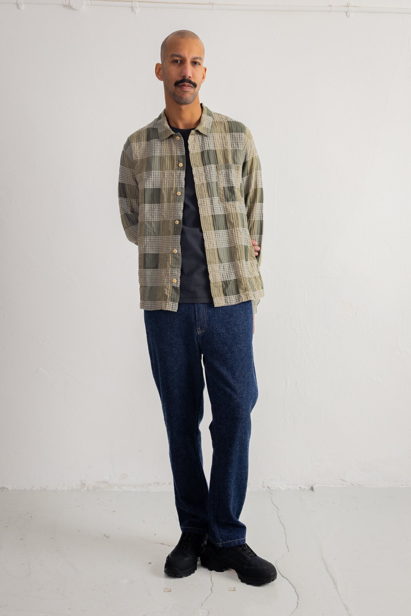 A man wearing a green and beige plaid button-up shirt over a dark t-shirt, paired with dark blue jeans and black shoes, standing against a white studio wall.