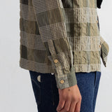 A close-up of a person wearing a long-sleeve plaid button-up shirt in olive green, beige, and gray tones, paired with dark blue jeans against a plain white wall.