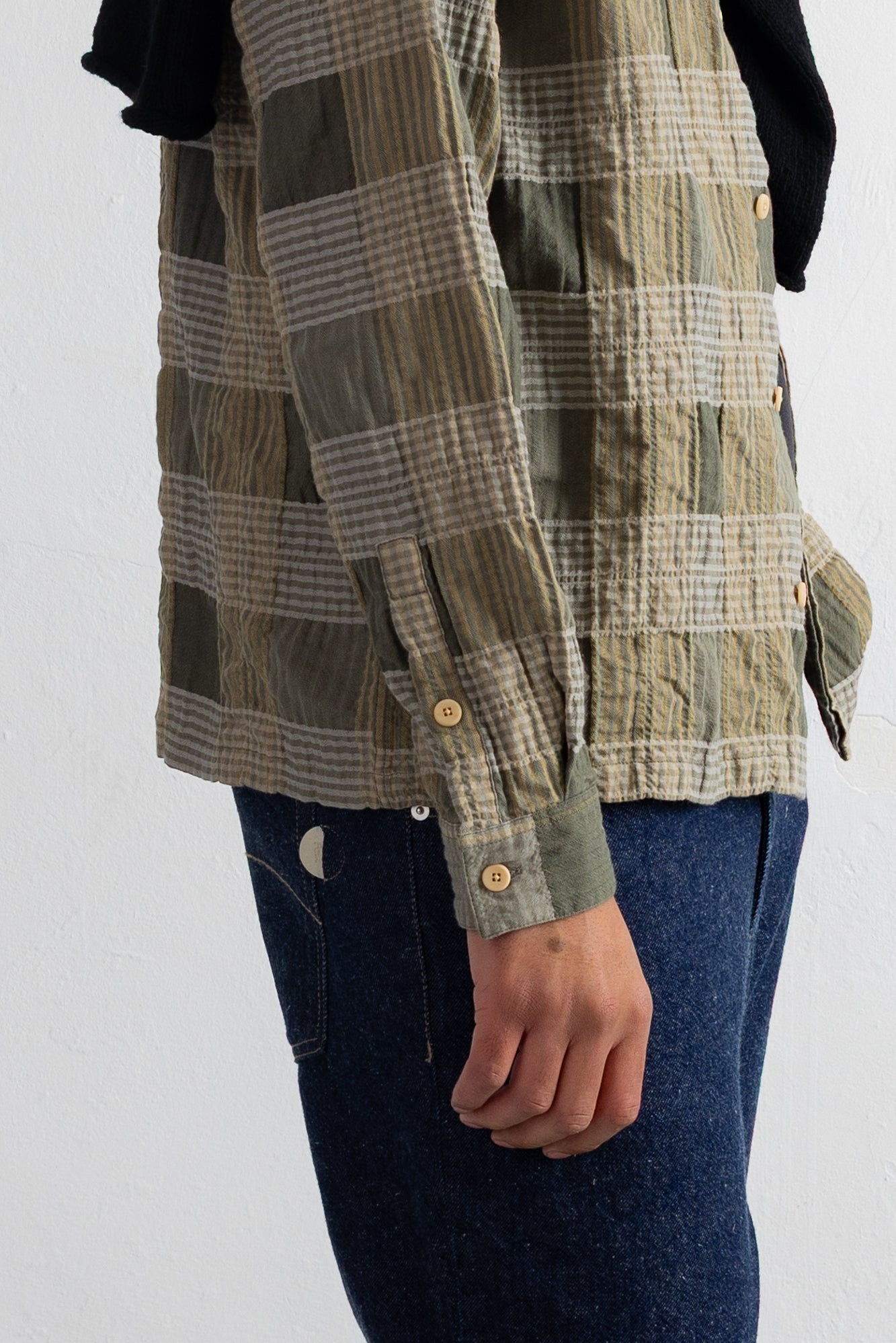 A close-up of a person wearing a long-sleeve plaid button-up shirt in olive green, beige, and gray tones, paired with dark blue jeans against a plain white wall.