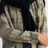A close-up of a person wearing a black sweater draped over an olive green and beige plaid button-up shirt, paired with a dark undershirt.