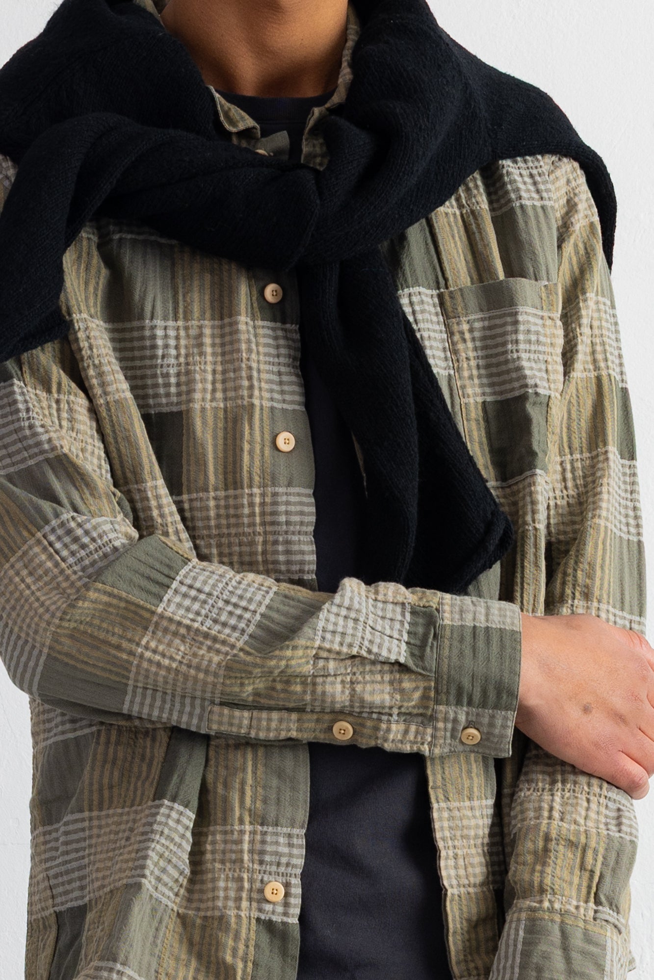 A close-up of a person wearing a black sweater draped over an olive green and beige plaid button-up shirt, paired with a dark undershirt.