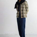 A man stands in a studio wearing a beige and brown plaid overshirt with a black knitted sweater draped around his neck, dark blue jeans, and black shoes.