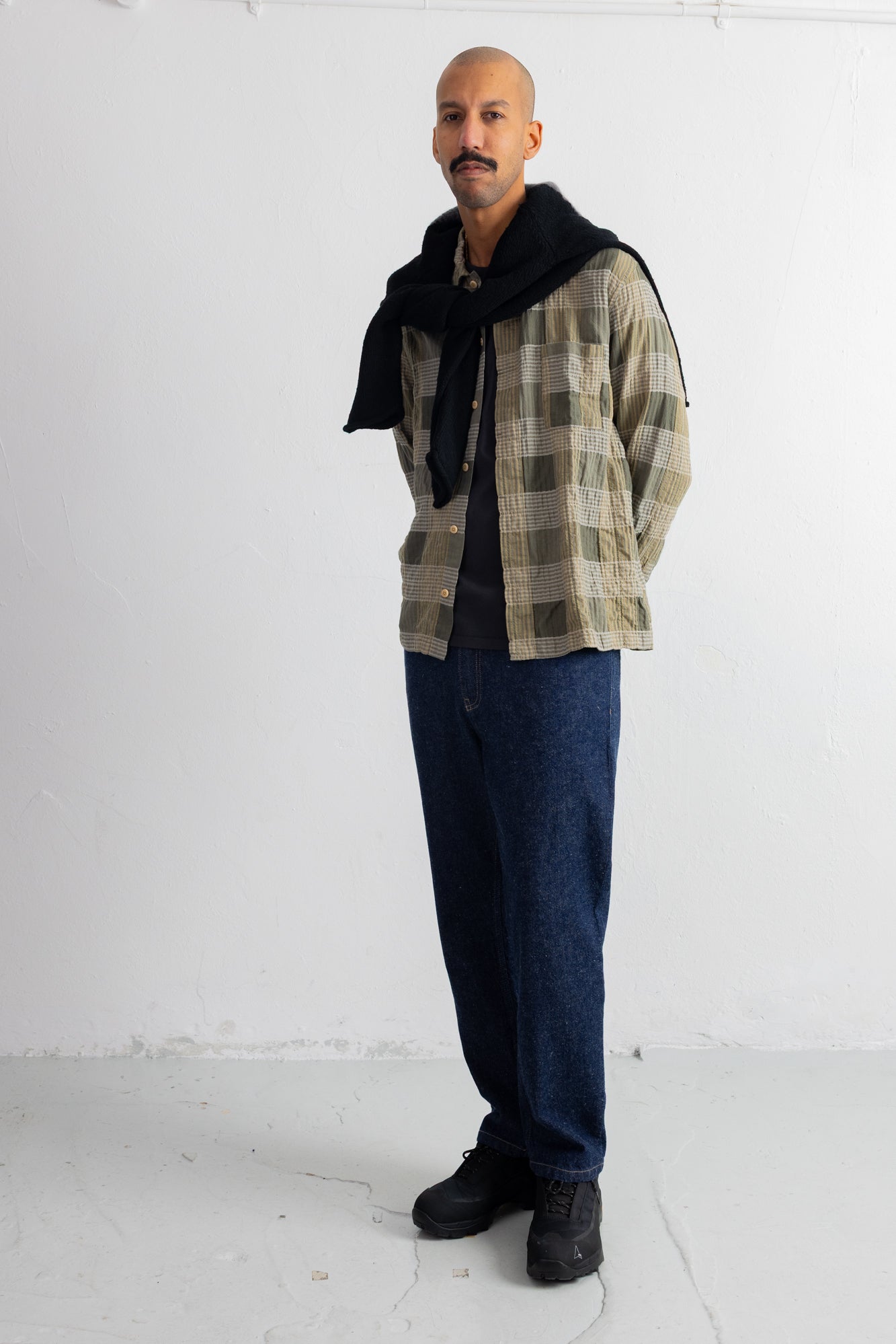 A man stands in a studio wearing a beige and brown plaid overshirt with a black knitted sweater draped around his neck, dark blue jeans, and black shoes.