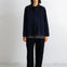 A model stands against a white studio wall wearing a dark navy corduroy jacket over a gray shirt, paired with dark charcoal trousers and black flats.