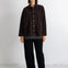 A model stands against a white studio wall wearing a dark brown corduroy button-up jacket with front pockets, black wide-leg trousers, and black pointed flats.
