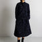 A model stands in a studio wearing a dark navy blue patterned quilted coat with a matching beanie and black boots.